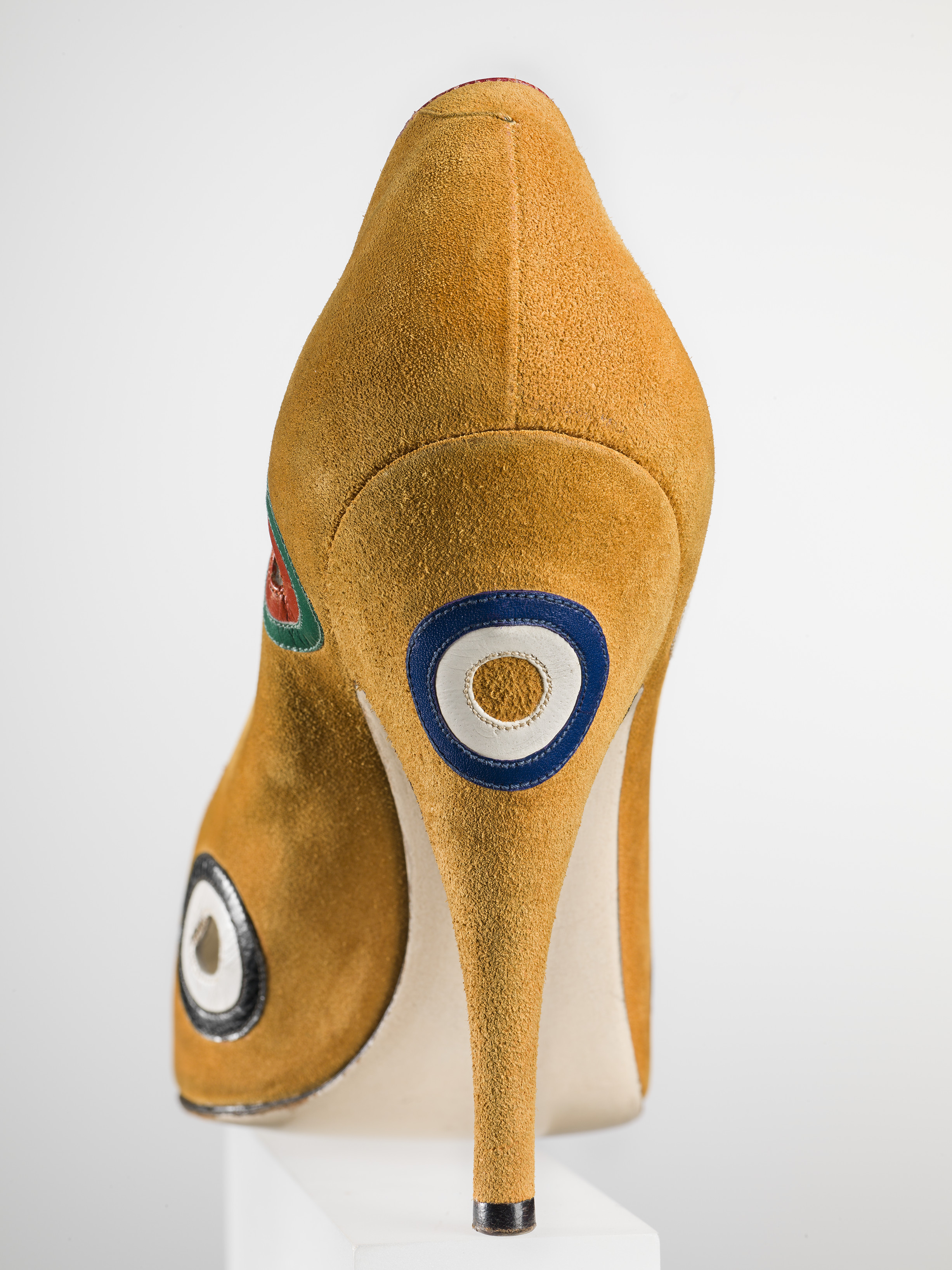 Salvatore Ferragamo, “Tirassegno” décolleté,1958, camoscio con applicazioni in capretto a motivo di tirassegno. Firenze, Museo Salvatore Ferragamo. Foto Arrigo Coppitz. Nel 1958 Ferragamo sul modello di scarpa realizzato per l’attrice Marilyn Monroe, crea un nuovo décolleté dal nome “Tirassegno”, che prende ispirazione dai dipinti creati nello stesso periodo dall’artista americano Kenneth Noland, studi su forme concentriche, come bersagli che con una sapiente combinazione di colori catturano l’occhio quasi ipnoticamente dando l’impressione di muoversi.